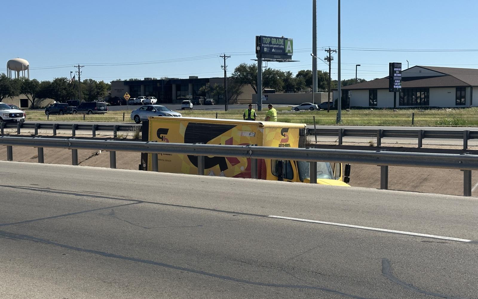 First responders assess the scene of a rollover involving a yellow delivery truck on W Loop 306. The vehicle, which had been flipped back onto its wheels, came to rest near a damaged guardrail. No injuries were reported.